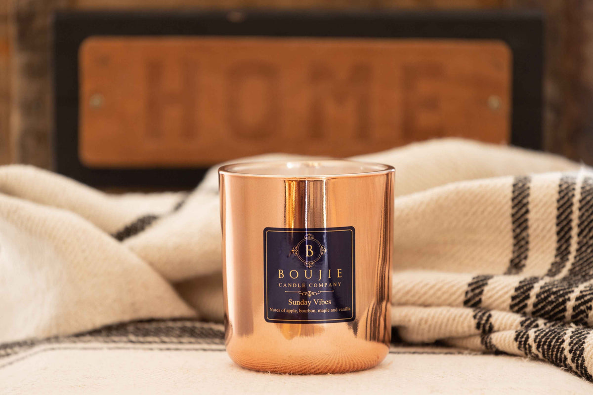 A Boujie Candle Company 'Sunday Vibes' scented candle in a reflective copper-colored glass jar, styled on a cream blanket with a black and white plaid throw and a blurred wooden "HOME" sign in the background.