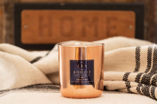 A Boujie Candle Company 'Sunday Vibes' scented candle in a reflective copper-colored glass jar, styled on a cream blanket with a black and white plaid throw and a blurred wooden "HOME" sign in the background.