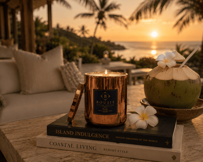 A lit copper glass vessel labeled Boujie Escape sits on stacked books on a table, with a coconut drink and white flowers in a tropical resort setting at sunset.