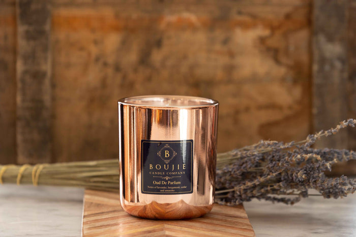 A copper-colored glass candle jar with a black square label reading Boujie Candle Company and Oud De Parfum, placed on a wooden board with dried lavender and a rustic wood backdrop.